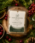 Clear plastic clamshell package of Cranberry Cedar Wax Melts by Naturehouse Wax Company, surrounded by fresh cranberries, cedar branches, and pine cones on a wooden surface.